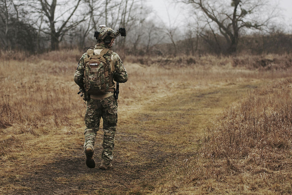 A soldier in camouflage walks on a dirt path through a dry, brown field. Bare trees are visible in the overcast background.