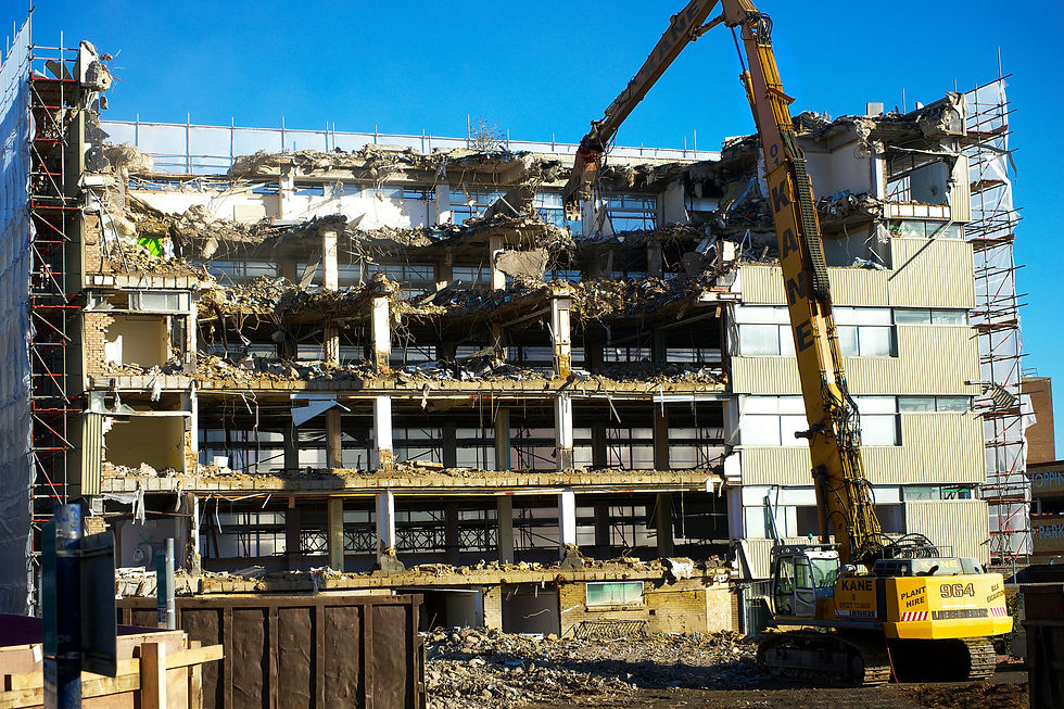 A demolition excavator tears down a multi-story building under a clear blue sky. Scaffolding and debris surround the structure.