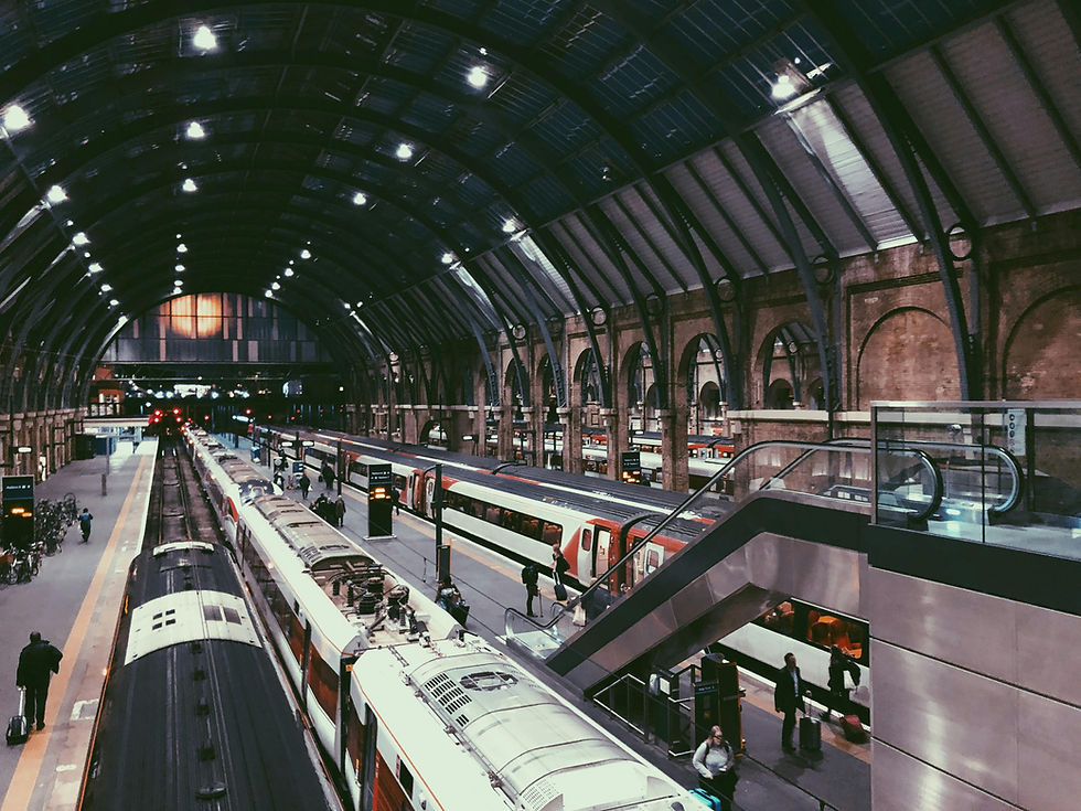 Train station interior with arched roof, platforms, and escalators. Trains and passengers visible, with warm lighting creating a busy atmosphere.