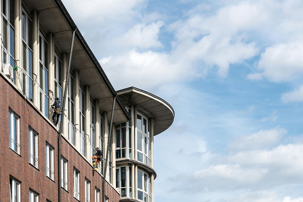 Window cleaners in harnesses work on a modern brick building with large windows. Blue sky with clouds in the background.