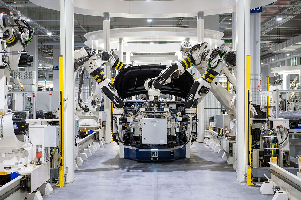 Robotic arms assemble a car on a factory line. Bright lights illuminate the industrial setting. Black and white machinery surrounds the vehicle.