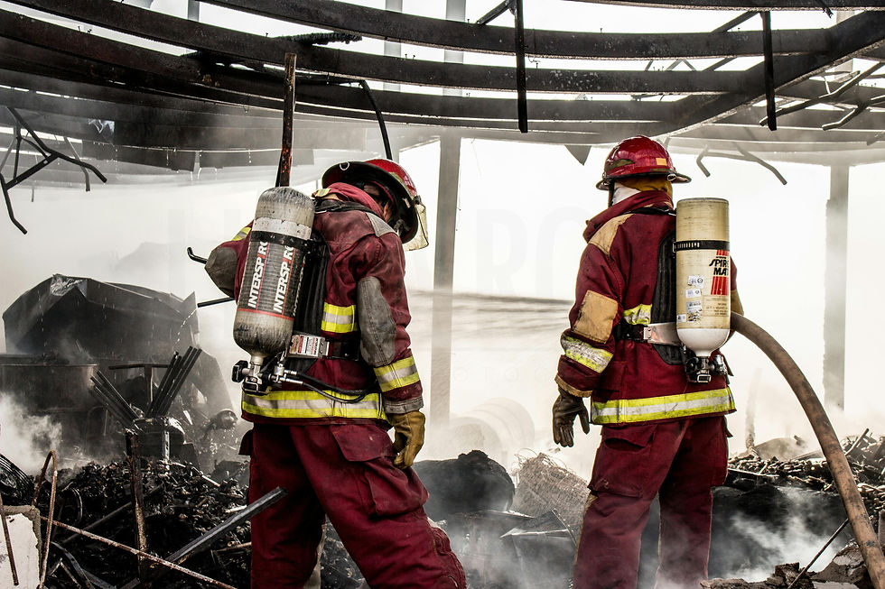 Two firefighters in full protective gear and breathing apparatus stand amidst smoke and debris inside a burned-out building, spraying water to extinguish remaining flames under a partially collapsed metal roof.