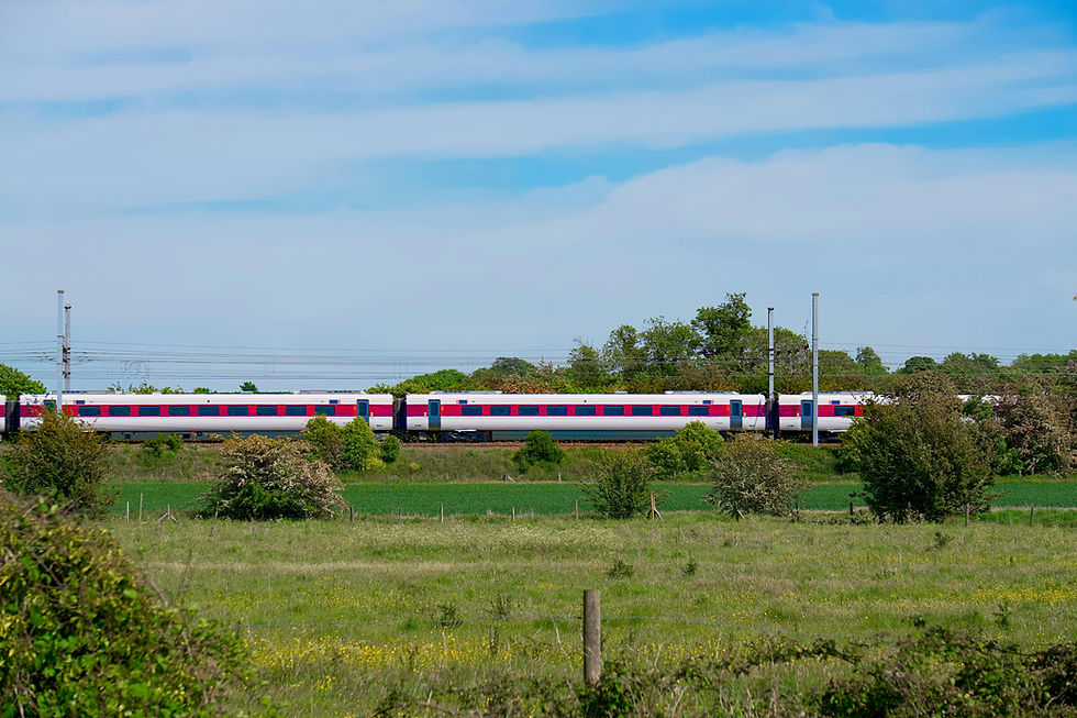 Train with red and white carriages passes through lush countryside with green fields and trees under a partly cloudy blue sky.