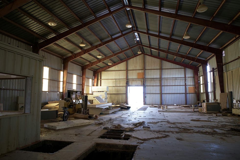 Spacious, dimly lit warehouse interior with stacked wood, machinery, and debris. Metal walls and roof, sunlight entering through an open door.