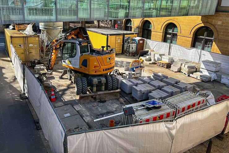 Construction site with a yellow excavator, stacks of stone slabs, and barriers. Yellow and brown walls in the background. Sunny day.