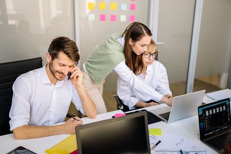 Three coworkers in a glass office work at laptops and phones, with colorful sticky notes on the wall, focused and collaborative.